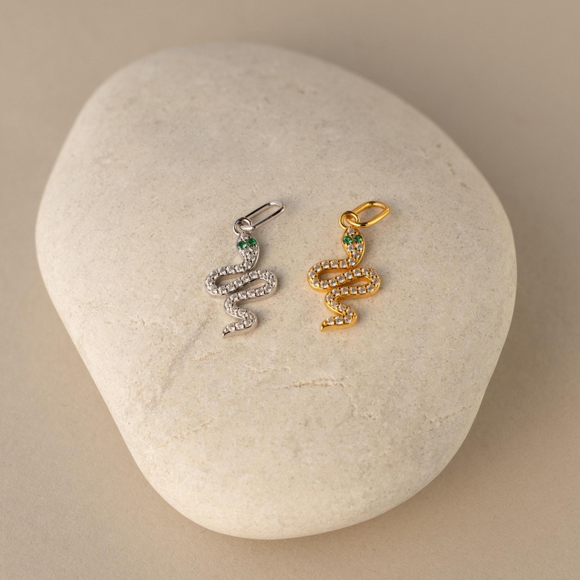 Two diamond serpent charm necklaces, one with a gold finish and the other with a silver finish, displayed on a textured stone surface.
